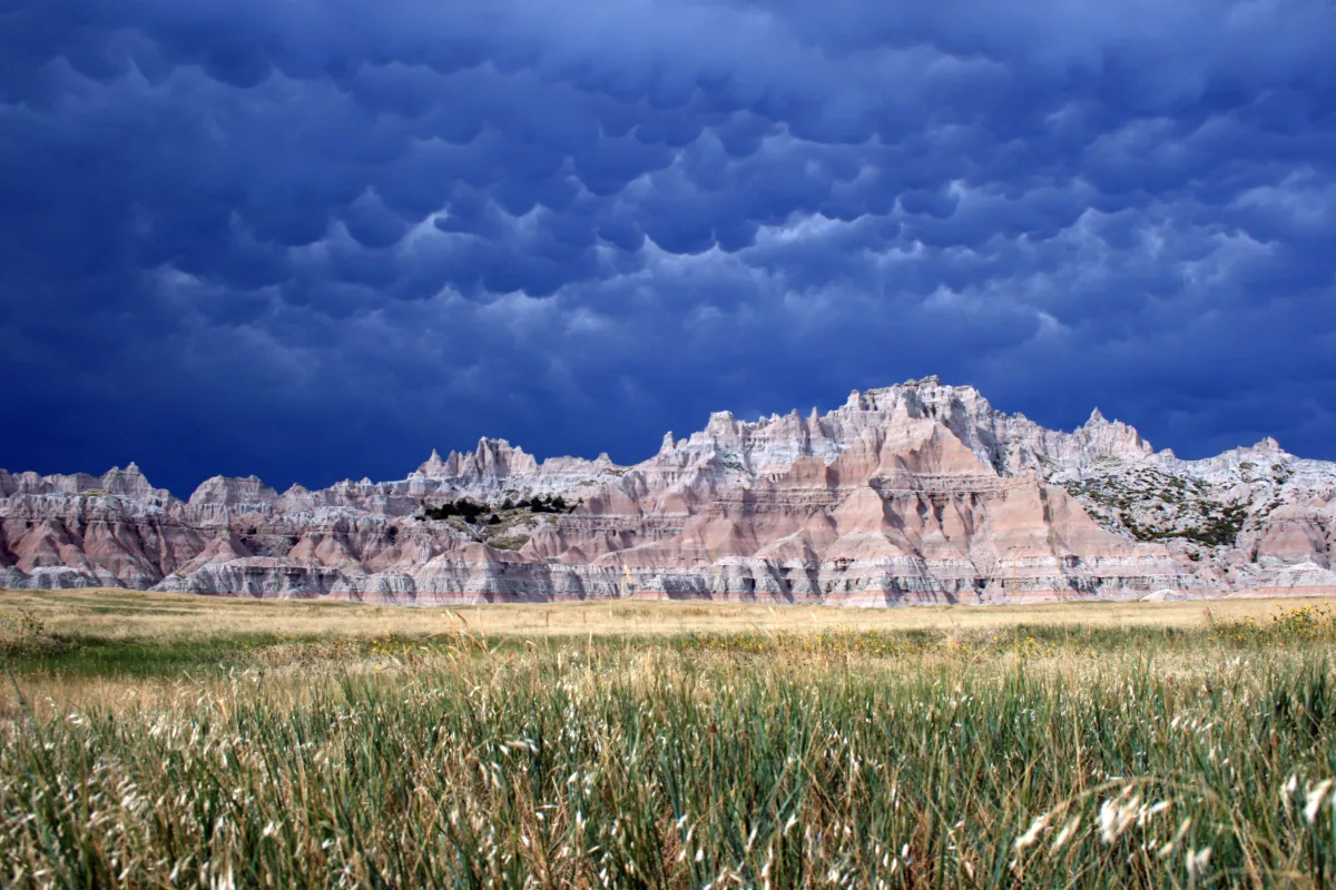 Layered badlands formations behind fields of green grass under cloudy and billowing clouds.
