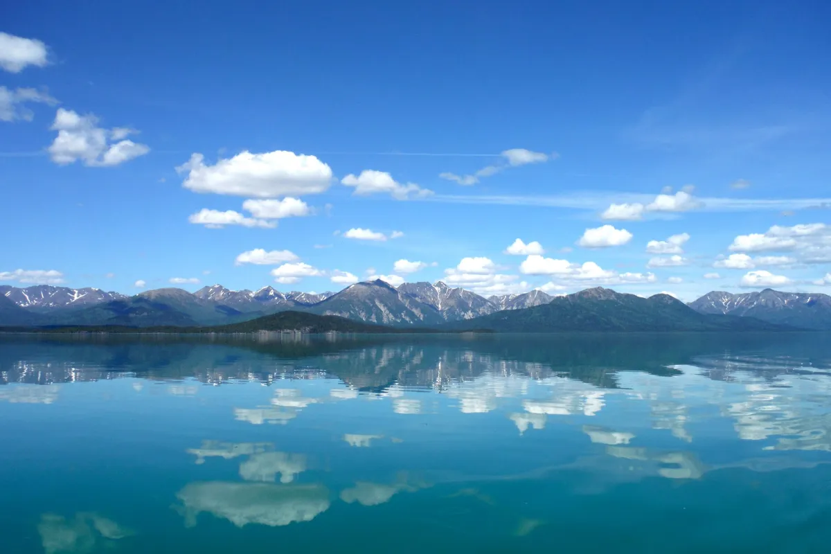 Photo of blue sky with fluffy white clouds reflect in calm lake with mountains in the background.