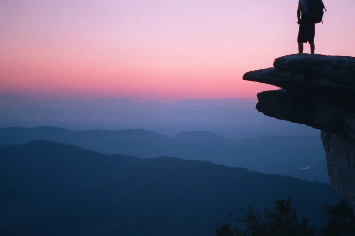 Silhouette of a man with backpack standing on McAfee Knob at sunset with mountains in the distance.