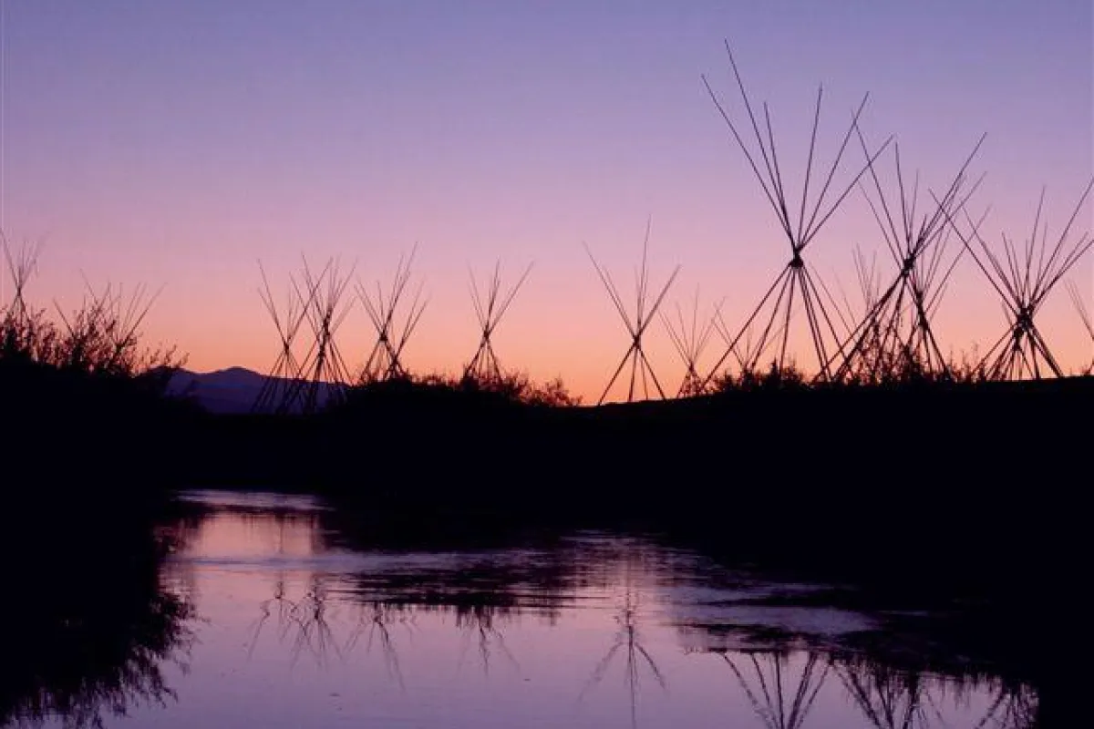Multiple tepee poles and a river are silhouetted against a dawn sky.