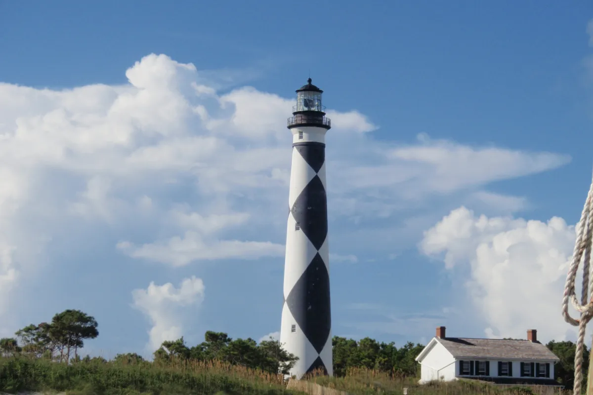 : Black & white patterned tower of the Cape Lookout Lighthouse stands against a blue & white cloud f