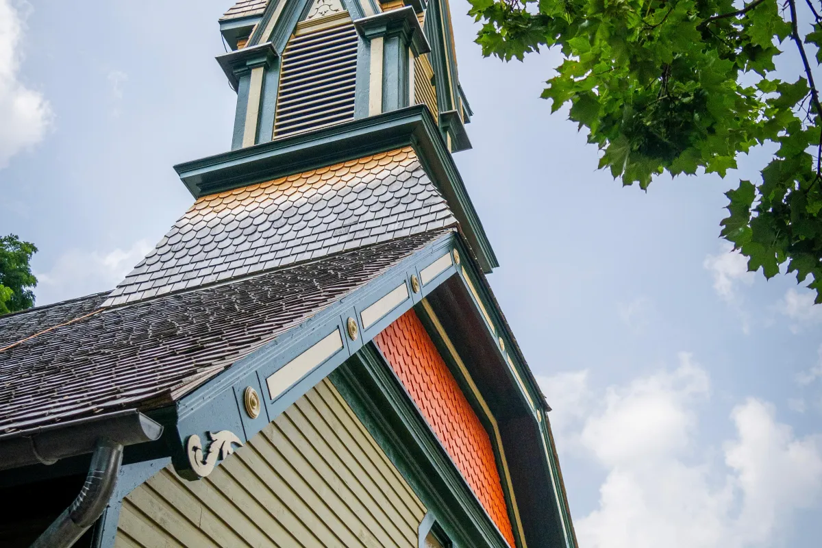 A tall, green and orange painted church steeple, against a blue sky, with a tree in the foreground.