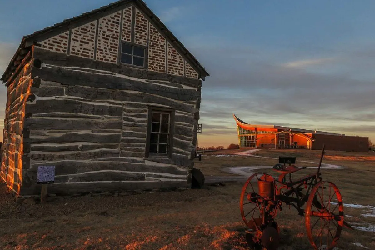 A historic cabin stands next to a concrete path that leads to a plow shaped visitor center