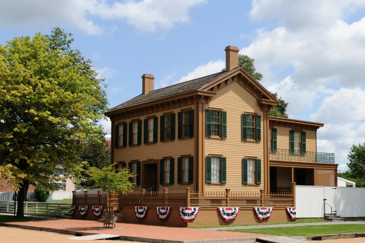 2 story tan house with green shutters. Red, white, and blue striped bunting draped below its fence