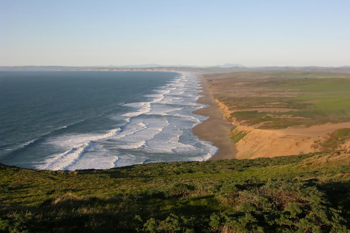 Waves wash in from the left onto a long, straight stretch of undeveloped, low-lying coastline.