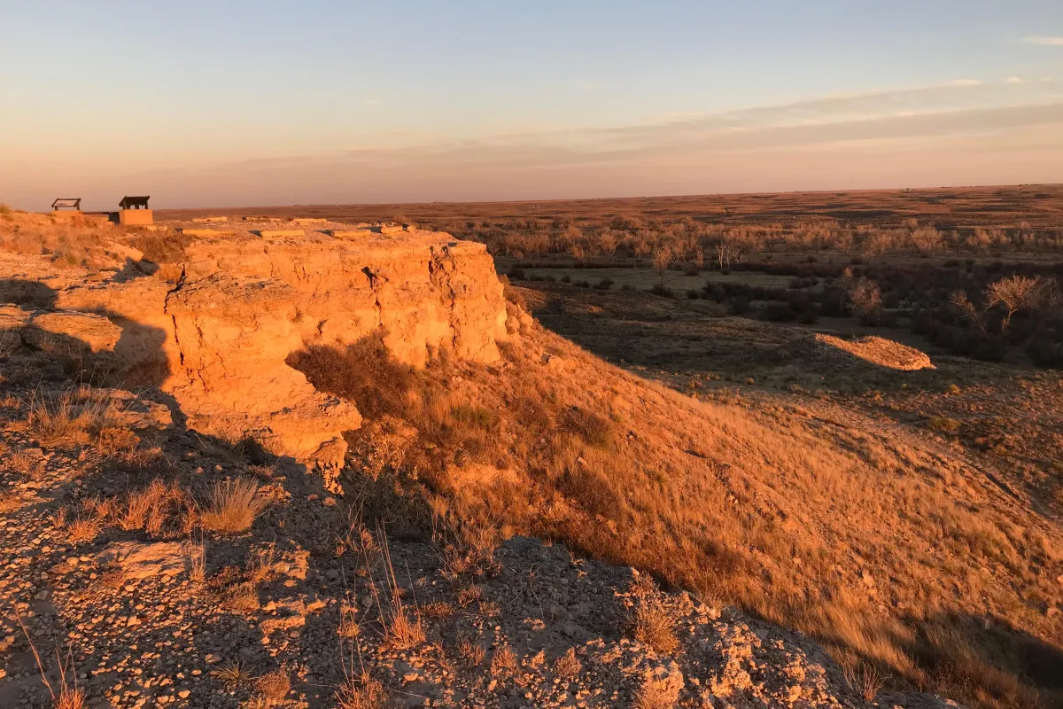 Warm light from the setting sun gives an orange glow to a rock bluff overlooking a grassland.