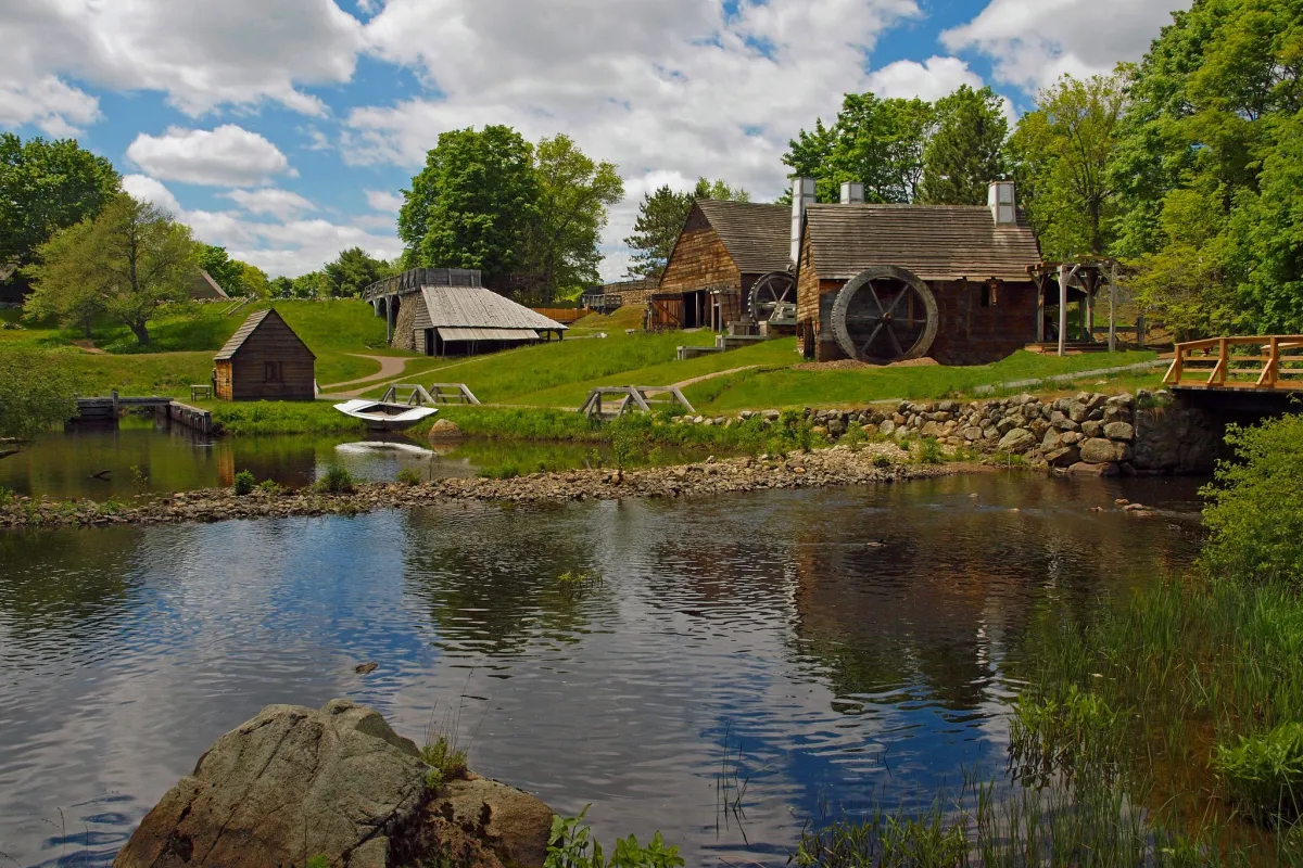 Several wooden structures amid green fields and trees under partly cloudy sky and beside river.