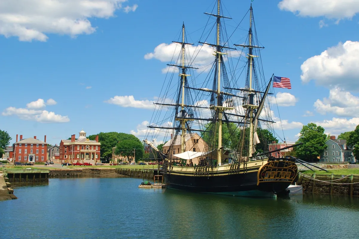 A three mast tall ship on the water under a blue sky with red brick buildings on the shore.