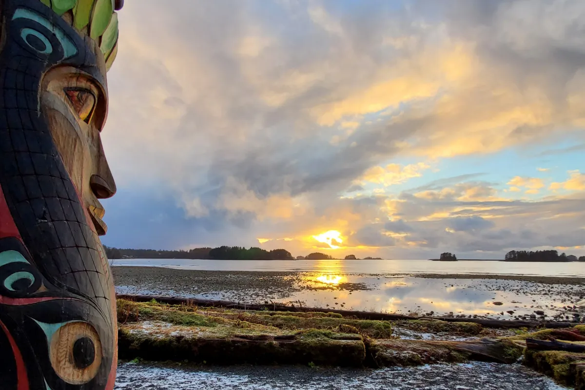 A totem pole in the foreground with a sunset over water in the background.
