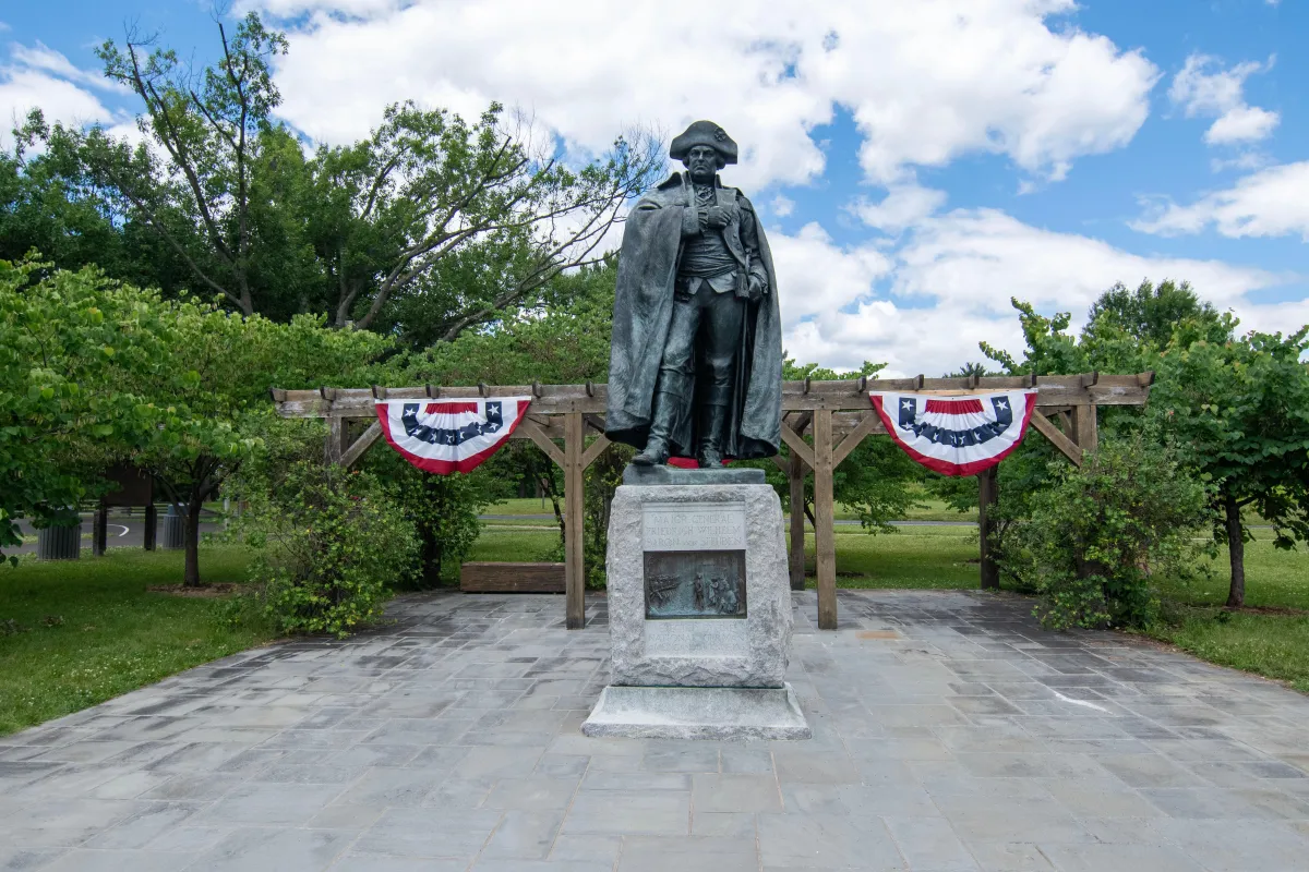 bronze statue of rev war general on stone base, red white and blue bunting behind